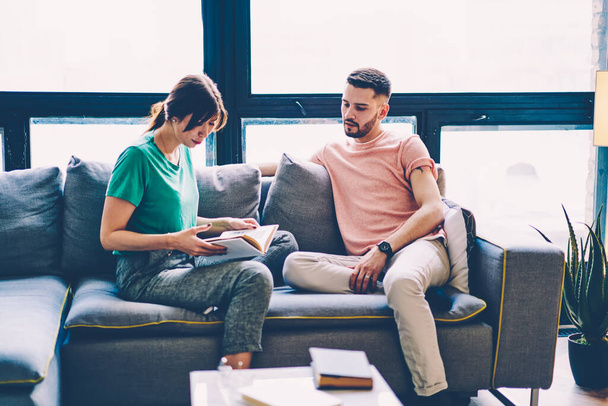 Young hipster couple sitting on sofa together discussing interesting literature on free time in home interior, beautiful woman holding book communicating with husband during leisure in living room - Photo, Image