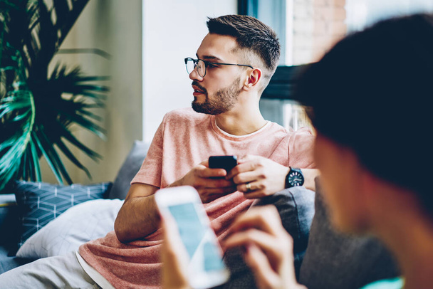 Young caucasian hipster guy in eyewear looking away using mobile phone for chatting sitting next to girlfriend at home, cropped image with blurred frontage of woman millennial holding smartphone - Zdjęcie, obraz