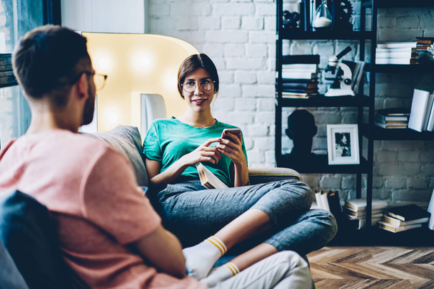 Cheerful caucasian woman in eyewear looking at her husband sitting on sofa with mobile phone on free time, smiling hipster girl enjoying resting at home with boyfriend using smartphone for browsing - Photo, Image