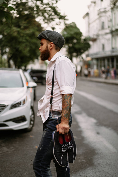 Young bearded tattooed man. A romantic guy in a white shirt, cap and suspenders walks in the city. revolves around a lamppost. Peaky Blinders. old-fashioned, retro. - Photo, Image