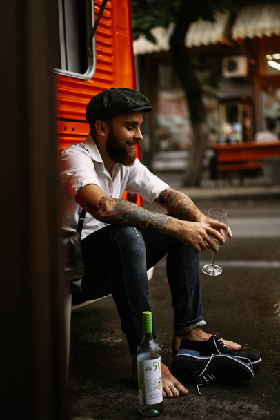 Young bearded tattooed man. A romantic guy in a white shirt, cap and suspenders walks in the city. revolves around a lamppost. Peaky Blinders. old-fashioned, retro. - Foto, Imagen