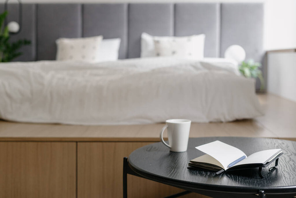 Εσωτερικό υπνοδωμάτιο, λευκό κρεβάτι διακόσμηση. Άνετο δωμάτιο με σκανδιναβικό σχεδιασμό, closeup στο τραπεζάκι του καφέ κοντά στο λευκό κρεβάτι. Έπιπλα για άνετη χαλάρωση, διαμέρισμα με ξύλινο αντικείμενο. - Φωτογραφία, εικόνα