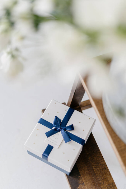 Vue du dessus du cadeau adressé à un être cher, ruban bleu avec arc, étiquette blanche avec des mots chaleureux écrits dessus, boîte posée sur l'échelle en bois, fleurs blanches floues dans un vase en verre. Plan vertical - Photo, image