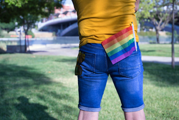 Reifer erwachsener Mann in einem senffarbenen T-Shirt und kurzen Jeans mit Sonnenbrille in der einen Gesäßtasche und Gay Pride Flagge in der anderen. - Foto, Bild