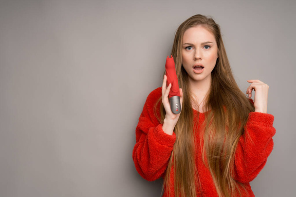 A white-skinned girl with long hair in a red jacket looks surprised holding a vibrator or a dildo for sex in her hand - Foto, afbeelding