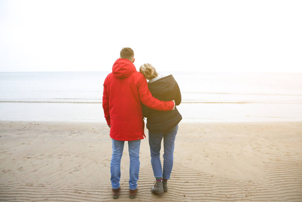 Two hipsters standing on the cold beach. Couple hugging and holding hands. Love story near the ocean. Winter season on the sea. Stylish boots on the sand. Man in red jacket. - Photo, Image