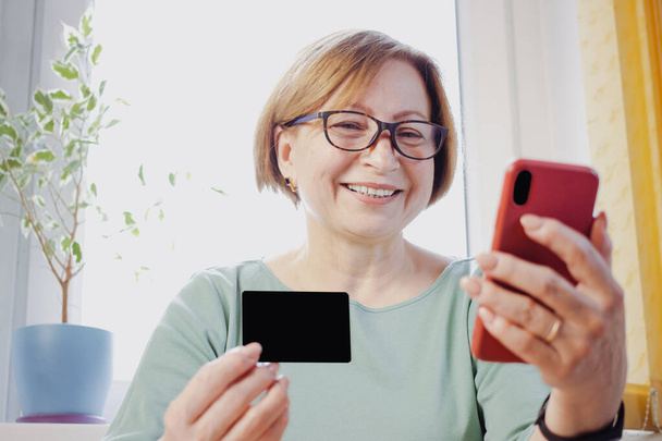 Senior blondi nainen ostoksia verkossa älypuhelimen ja pankkikortilla, istuu kotona. Happy 50s vanhukset nainen oppimisen maksujärjestelmä, valitsemalla palvelu, tuote verkkokaupassa, varaus hotelli - Valokuva, kuva