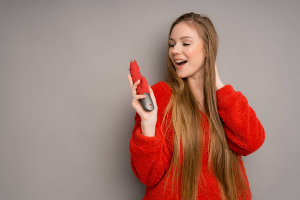 An attractive girl of European appearance in a red sweater happily holds a red phallus - Zdjęcie, obraz