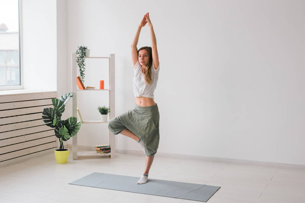 Woman practise yoga performs. Tree pose standing on mat inside of cozy room with plants and greenery. Healthy lifestyle concept. Copy space. - Fotoğraf, Görsel