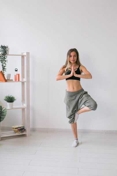Woman practise yoga performs. Tree pose standing on mat inside of cozy room with plants and greenery. Healthy lifestyle concept - Foto, Bild