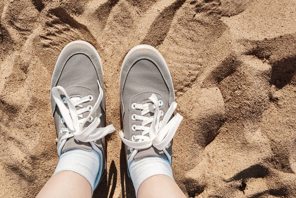 Legs in gray sneakers on the beach sand. Summer vacation concept by the sea. Lifestyle travel. Pov view. Copy space. - Foto, afbeelding