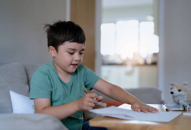 Az iskolás fiú ceruzarajzot használ fehér papírlapra, a fiatal srác iskolai házi feladatot csinál, a boldog vegyes fajú gyerek szeret kézművességet csinálni otthon a hétvégén. Otthoni oktatás  - Fotó, kép