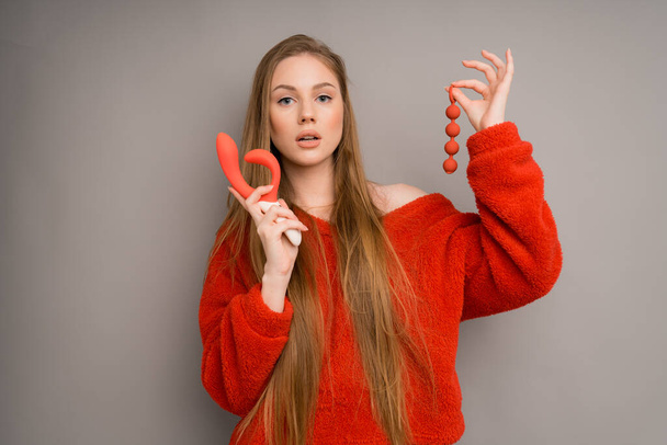 Attractive caucasian girl in a red sweater depicts the emotion of silence, puts her index finger to her lips and holds a red dildo and vaginal balls in her hand, a massager for sex - Foto, immagini
