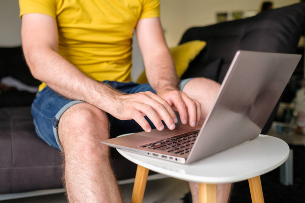 Man in het gele T-shirt typt op de laptop zittend in de moderne woonkamer. Freelancer werkt thuis - Foto, afbeelding