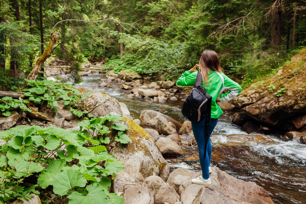 Traveler hiker with backpack enjoys landscape by mountain river in Carpathian forest. Woman tourist enjoys view. Trip to summer Ukraine - 写真・画像
