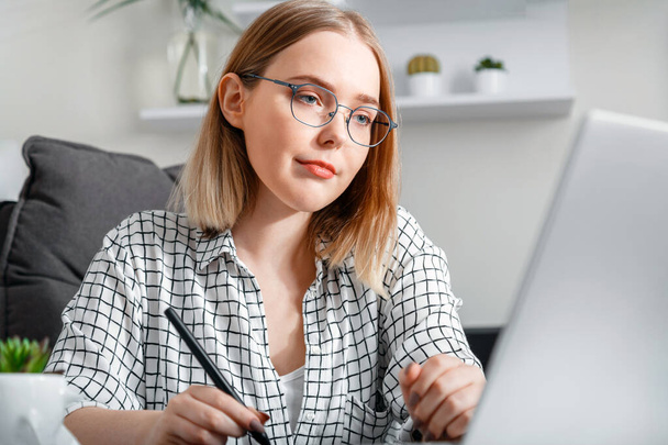 Confident blonde woman in glasses working using laptop while making notes in office interior. Student studying e learning at home. - Zdjęcie, obraz