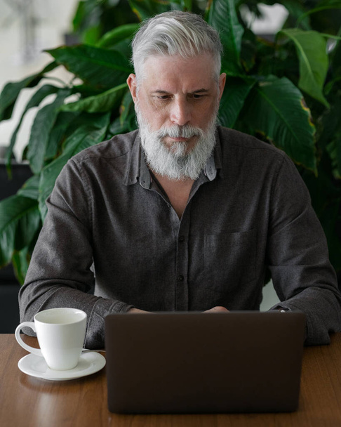 A grown man with gray hair and a beard works at his laptop in his office or airport waiting area and drinks coffee - Fotoğraf, Görsel