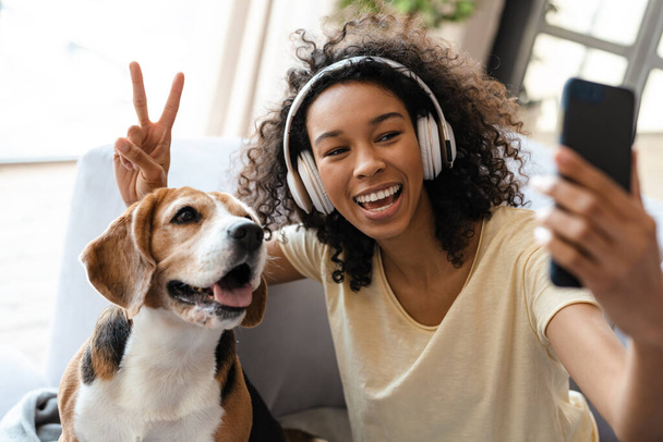 Happy young african woman in headphones relaxing on a couch with her pet dog at home, taing a selfie - Fotografie, Obrázek