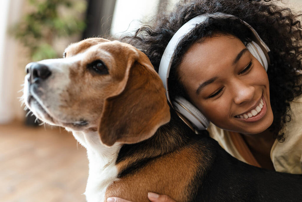 Happy young african woman in headphones relaxing on a couch with her pet dog at home, cuddling - Foto, immagini