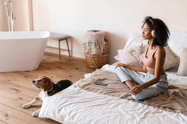 Young african woman in casual wear with beagle puppy at home, relaxing on bed in headphones, meditating - Photo, image