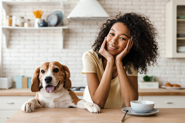 Smiling young african woman leaning on a kitchen counter with her pet beagle - Foto, afbeelding
