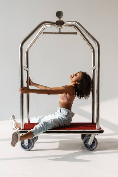 Fashion portrait of a young curly african woman posing on a luggage trolley over white wall backgroud, full length - Foto, imagen