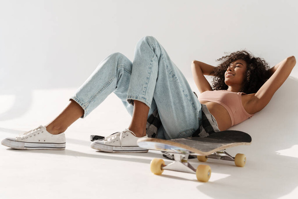 Young african woman smiling while lying with skateboard on floor isolated over white wall - Fotó, kép