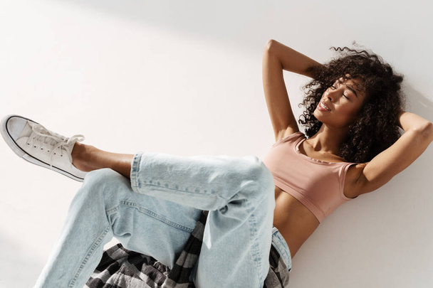 Young african woman wearing jeans smiling while lying on floor isolated over white wall - 写真・画像
