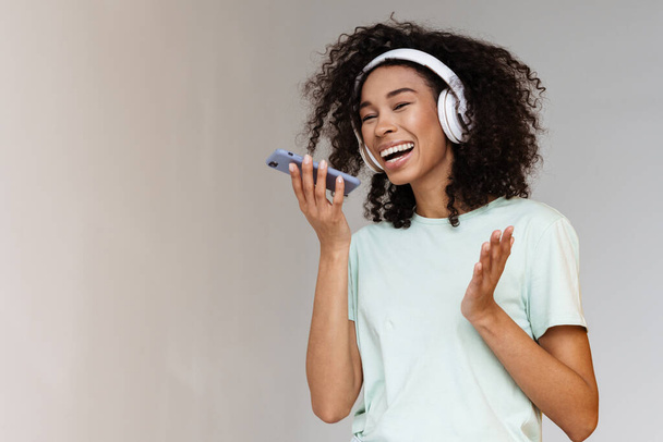 Young african woman in headphones smiling while using mobile phone isolated over white wall - Foto, immagini
