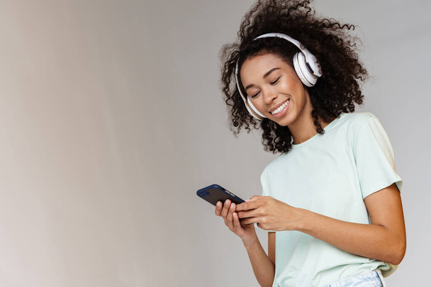 Young african woman in headphones smiling while using mobile phone isolated over white wall - Photo, Image