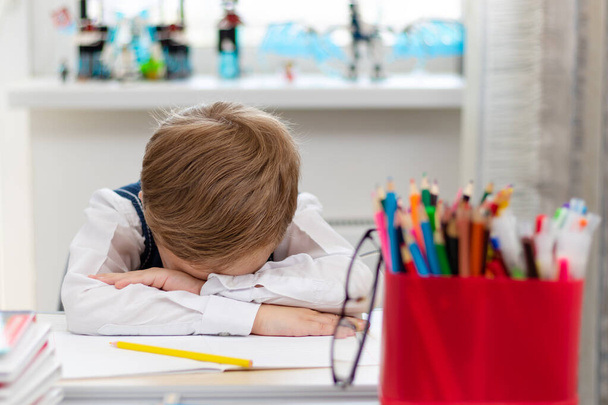 Egy cuki elsőéves fiú iskolai egyenruhában otthon, világjárvány idején elaludt házi feladatot csinálni egy íróasztalnál könyvekkel és ceruzákkal. Szelektív fókusz. Közelkép. Portré - Fotó, kép