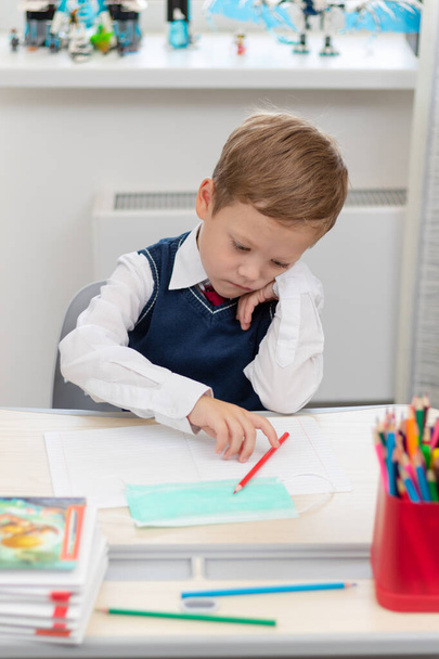 Ένα χαριτωμένο αγόρι της πρώτης δημοτικού με σχολική στολή κάνει τα μαθήματά του ενώ κάθεται σε ένα γραφείο με ένα μολύβι στα χέρια του στο σπίτι. Απομακρυσμένη εκπαίδευση. Επιλεκτική εστίαση. Κοντινό πλάνο. Πορτρέτο - Φωτογραφία, εικόνα