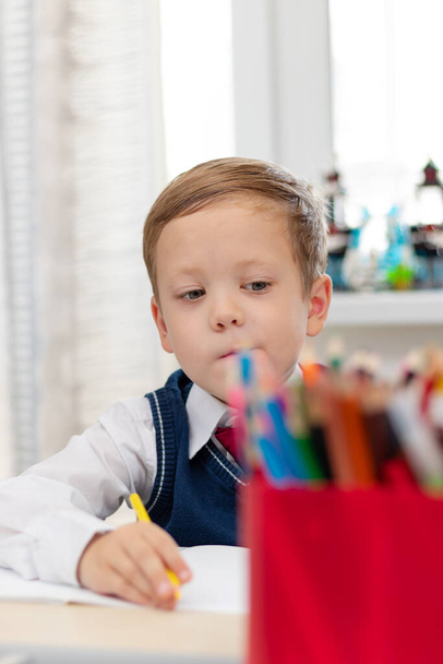 Egy cuki elsőéves iskolai egyenruhában házi feladatot csinál, miközben otthon ül egy íróasztalnál egy ceruzával a kezében. Távoktatás. Szelektív fókusz. Közelkép. Portré - Fotó, kép