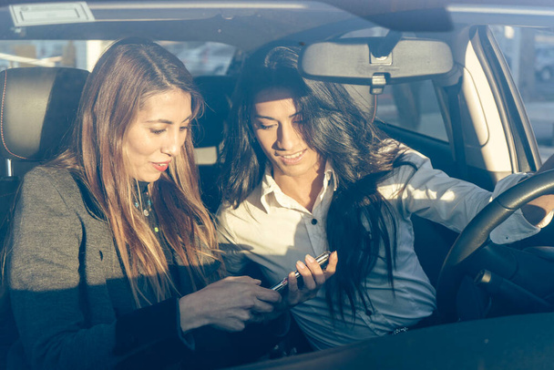 Two beautiful Latina women in a car listening to a message from a cell phone. Communications, technology concept. - Foto, imagen