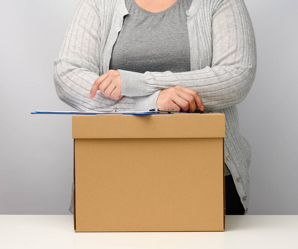 Женщина в серой одежде стоит возле коричневой коробки. Концепция перемещения, отправки и приема грузов. Увольнение с работы - Фото, изображение