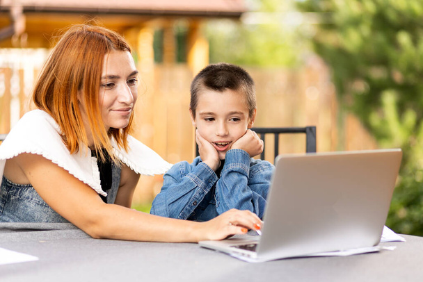 mom teaches the child at home in the garden, home schooling. Mom helps her son do his homework, stress. Online school lessons via laptop - Photo, Image