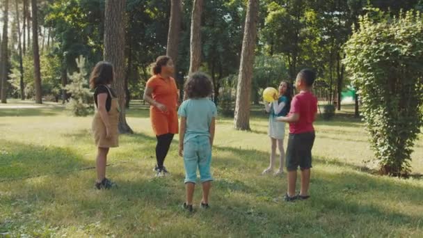 Školní děti hrát míč s mladou učitelkou ve veřejném parku - Záběry, video