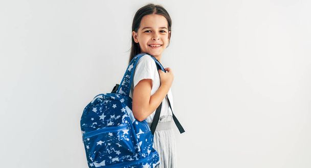 Side view of positive schoolgirl with blue backpack smiling and looking at camera. Pretty schoolkid with backpack has joyful expression. Studio portrait of little girl posing over grey background.  - Foto, imagen
