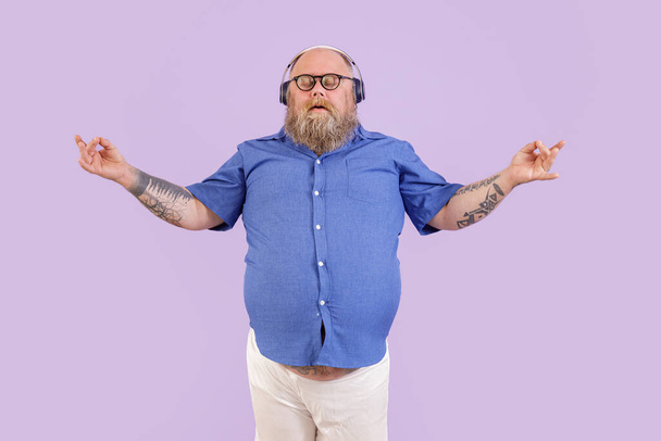 Tranquil obese man meditates listening to relaxing music on purple background - Photo, Image