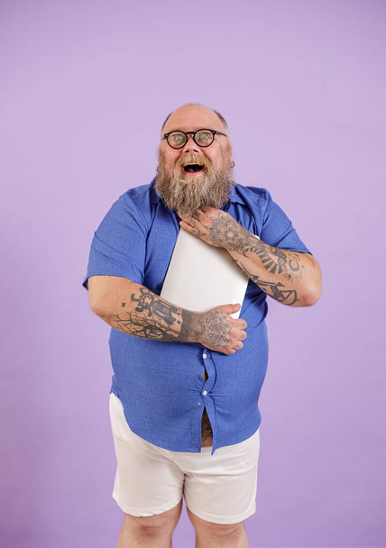 Ενθουσιασμένος γενειοφόρος παχύσαρκος άντρας με στενό πουκάμισο αγκαλιάζει μοντέρνο φορητό υπολογιστή σε μωβ φόντο - Φωτογραφία, εικόνα