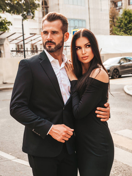 Beautiful fashion woman and her handsome elegant boyfriend in suit. Sexy brunette model in black evening dress. Fashionable couple posing in the street in Europe. Brutal man and his female outdoors - Foto, imagen