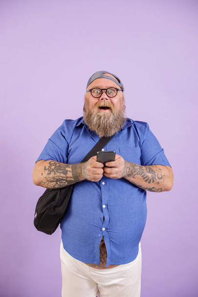 Vicces plusz méret férfi kereszttest táska és szemüveg tartja telefon lila háttér - Fotó, kép