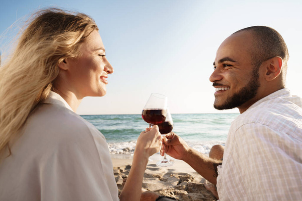 Portrait of a young couple sitting on the beach and drinking wine - Fotoğraf, Görsel