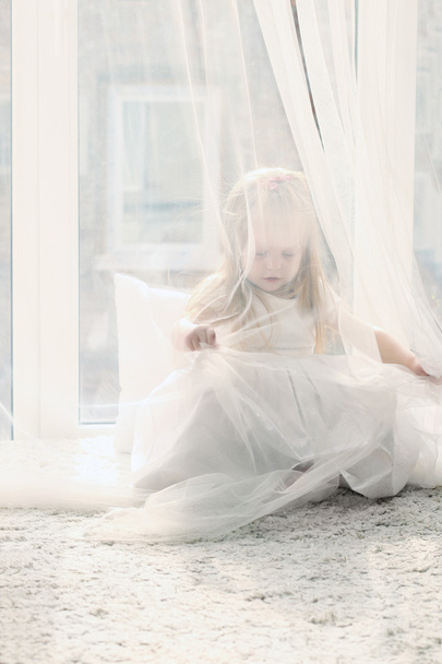Pretty little blonde girl sits near big window and hides behind  - Φωτογραφία, εικόνα