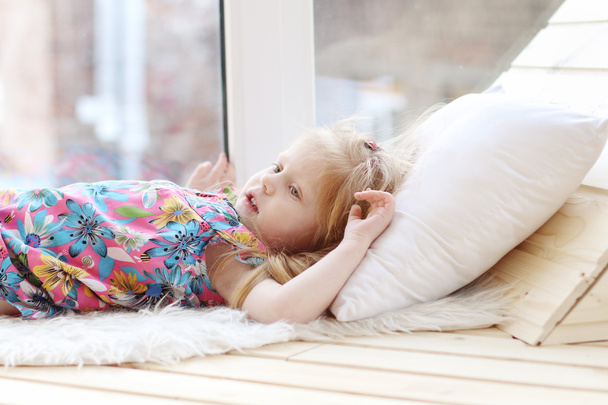 Pretty little blonde girl lies on white pillow near window in ro - Photo, image