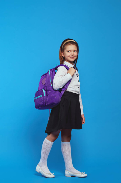 Adorable schoolgirl wearing uniform and backpack, smiling against blue wall - Foto, afbeelding
