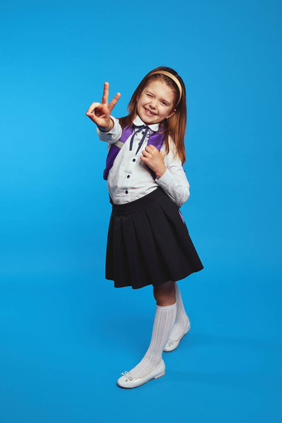 Schoolgirl wearing uniform and backpack smiling while showing peace gesture - Fotó, kép