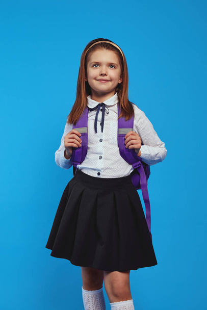 Adorable schoolgirl wearing uniform and backpack, smiling against blue wall - Valokuva, kuva