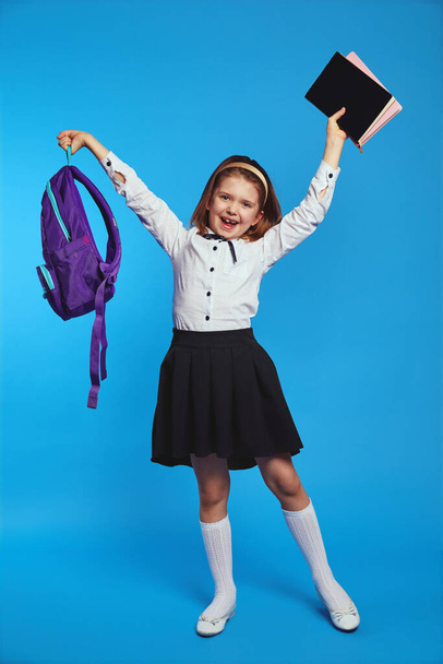 Школьница поднимает букет книг и рюкзак в руке и улыбается - Фото, изображение