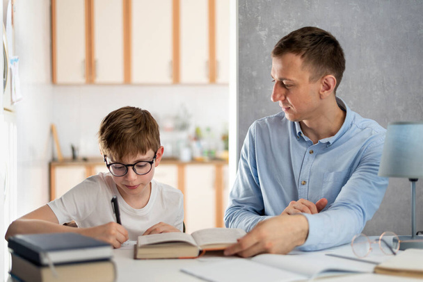 Padre e hijo están haciendo la tarea. El joven profesor tutor está estudiando con un chico. Niño adolescente con gafas escribe en un cuaderno - Foto, imagen